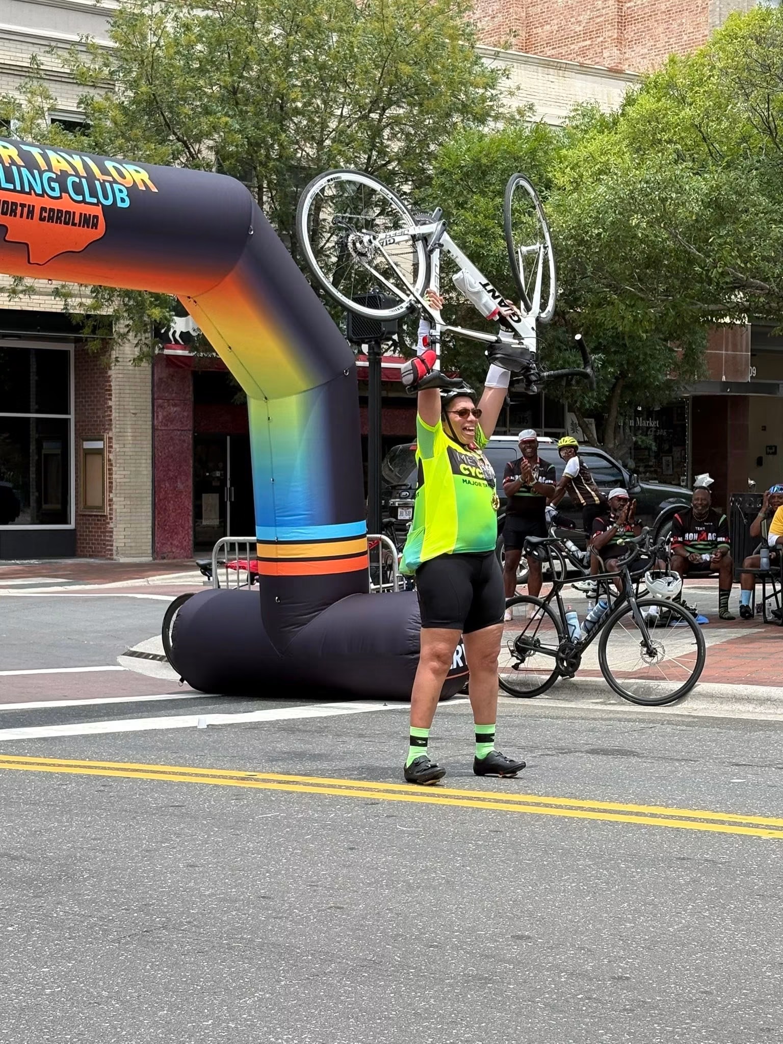 Cyclist raising her bike overhead in celebration at the finish line arch in downtown Durham at the MTICA 2026 Convention