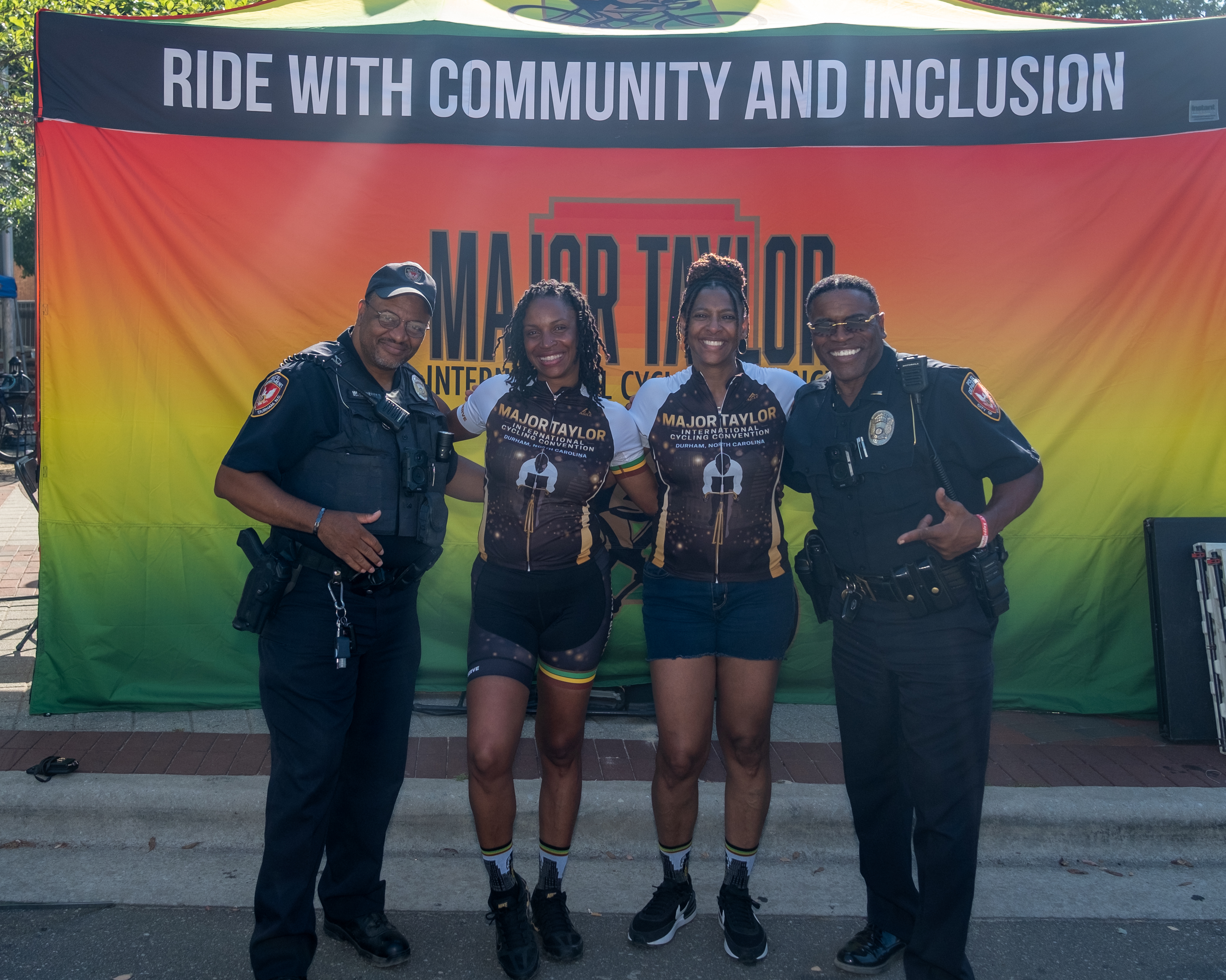 MTCCNC riders in Major Taylor International Cycling Convention jerseys posing with Durham Police Department officers at the Ride With Community and Inclusion banner